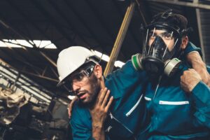 An employee wearing a gas mask and supporting a colleague who is semi-collapsed and has their hand up to their throat.