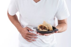 A man in a white t-shirt holding his stomach with a dumpling in his hand.