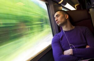 A man in a purple jumper sat on a train looking out the window with his arms crossed and wired earphones in. 