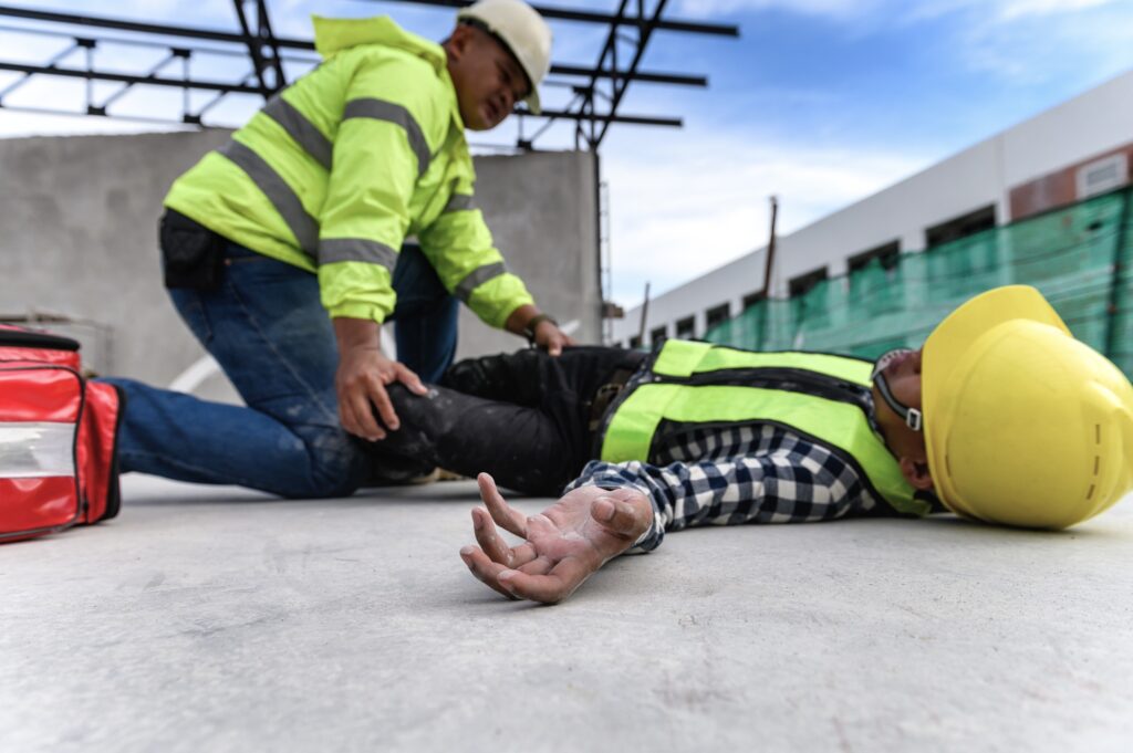 A worker laying on the ground after an accident at a construction site while his colleague provides first aid. 