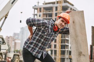 A construction worker in an orange hard hat. They are bent over and holding their back while grimacing.