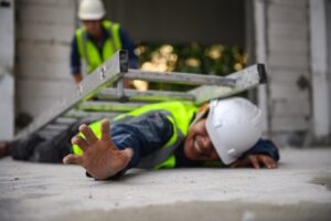 A person in a hi-viz jacket and a white helmet lies on the floor, face-down. There is a fallen ladder on top of them.