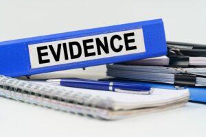 A blue ring binder labelled "evidence" sits on top of a small pile of notepads, documents, and stationary.