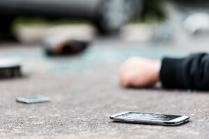 A shattered mobile phone and a person's arm lay in front of a blurred car in the background.