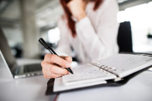 A woman sitting at a desk besides an open laptop uses a pen to take notes.